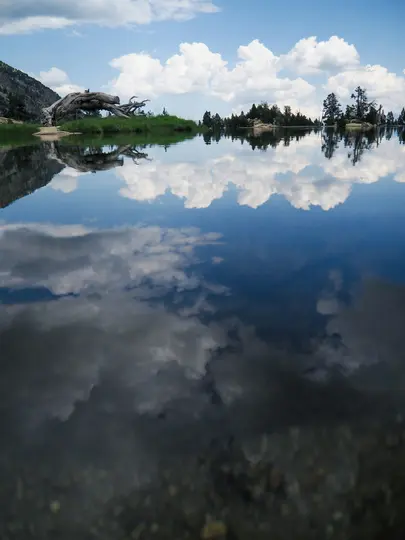 En stilla sjö reflekterar en blå himmel med fluffiga vita moln. Till vänster finns ett berg och en stor bit drivved…