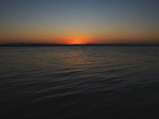 Ett brett landskapsfoto av en solnedgång över ett lugnt vattenområde. Solen är en ljus orange skiva vid horisonten och