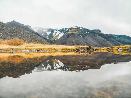 En vidsträckt landskapsvy över mörka, karga berg med snöfläckar, perfekt återspeglade i ett stilla vattenområde. Gyllene…