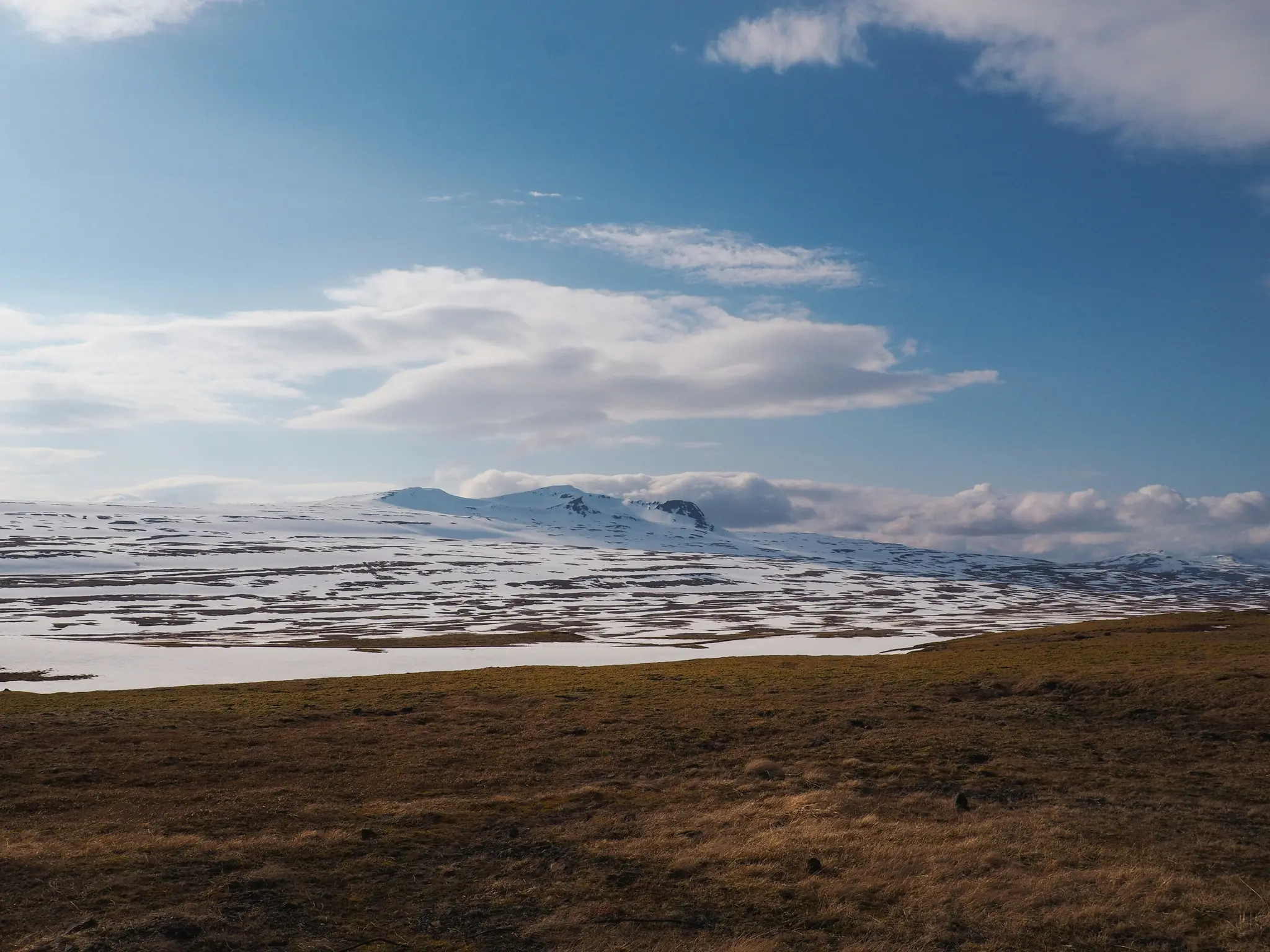 En vid landskapsvy över snötäckta berg och fält med fläckar av brunt och grönt gräs under en klarblå himmel…