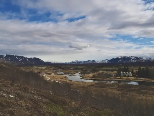 En bred bild av en dal med en slingrande flod som speglar himlen, omgiven av bruna och gröna fält. Snötäckta…