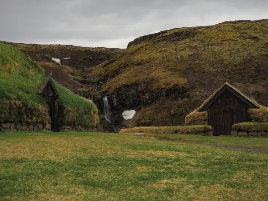 En vidvinkelbild av två traditionella hus med torvtak i ett frodigt grönt landskap. Ett vattenfall kaskaderar nerför en