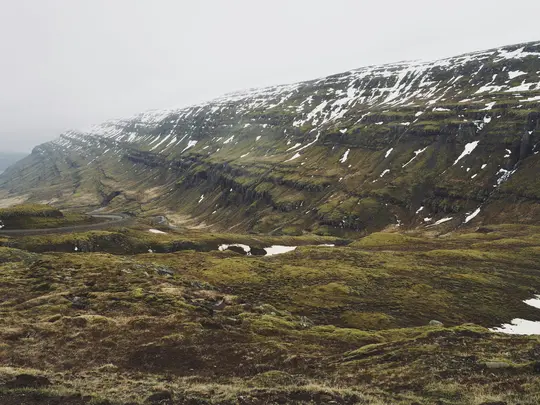 En bred landskapsvy av en slingrande väg som skär genom en dal. Bergssluttningarna är täckta av grön mossa och fläckar...