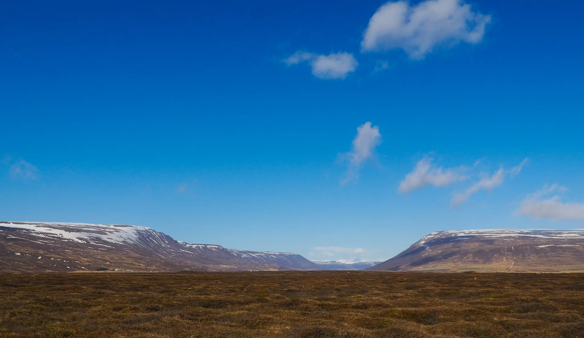 Ett brett landskapsfoto som visar ett stort, platt, brunt och grönt fält i förgrunden, som leder till snötäckta berg.
