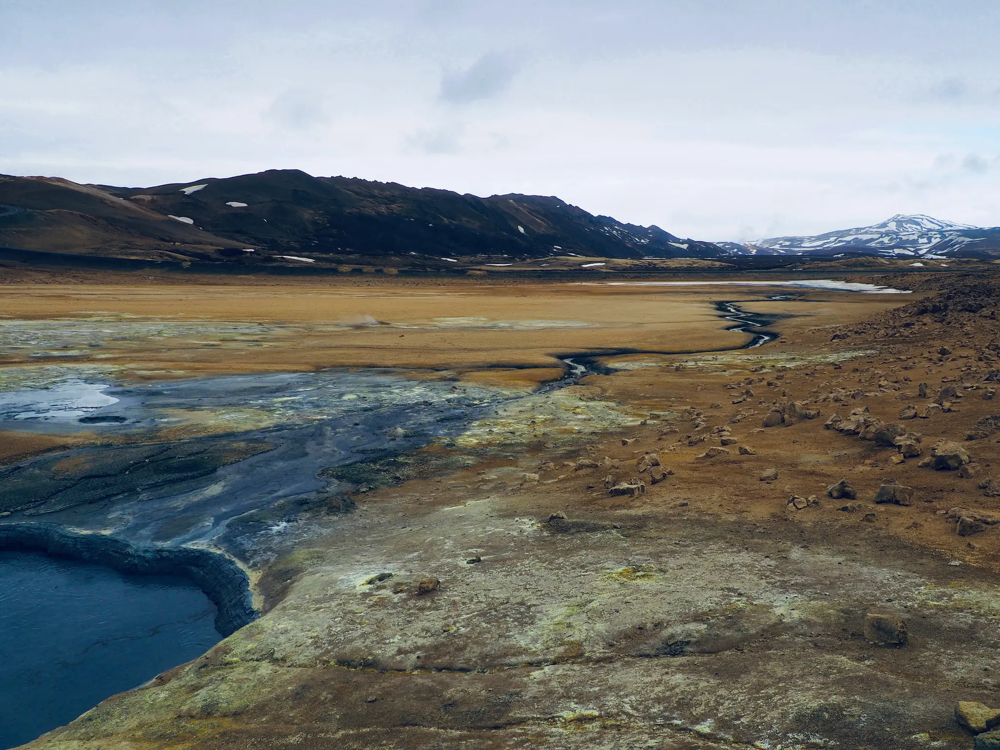 En vidvinkelbild av ett geotermiskt landskap med en mörkblå pool, färgglad mineralfärgad mark och en slingrande bäck.