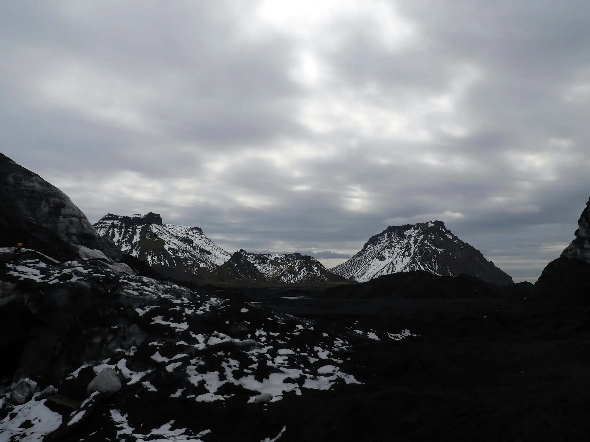 En vidvinkelbild av ett kargt, mörkt vulkaniskt landskap med flera snötäckta berg under en kraftigt molnig himmel. Landskapet