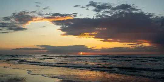 Laaja maisemakuva auringonlaskusta meren yllä. Aurinko on kirkkaan oranssi kiekko lähellä horisonttia, heijastuen…