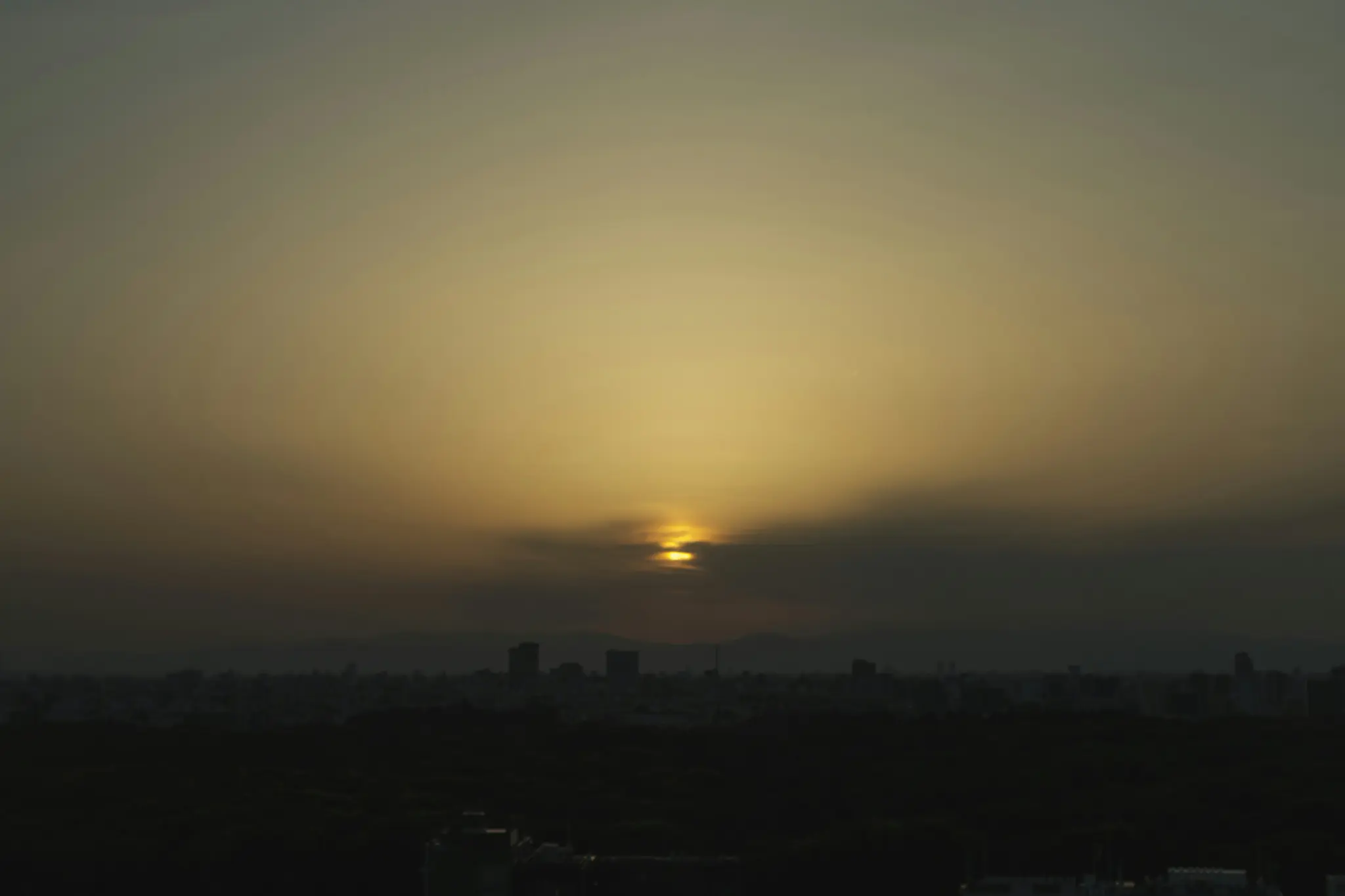 A wide landscape photo of a sunset over a city skyline, with the sun partially obscured by clouds and a dark foreground.