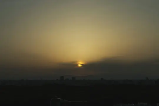 A wide landscape photo of a sunset over a city skyline, with the sun partially obscured by clouds and a dark foreground.
