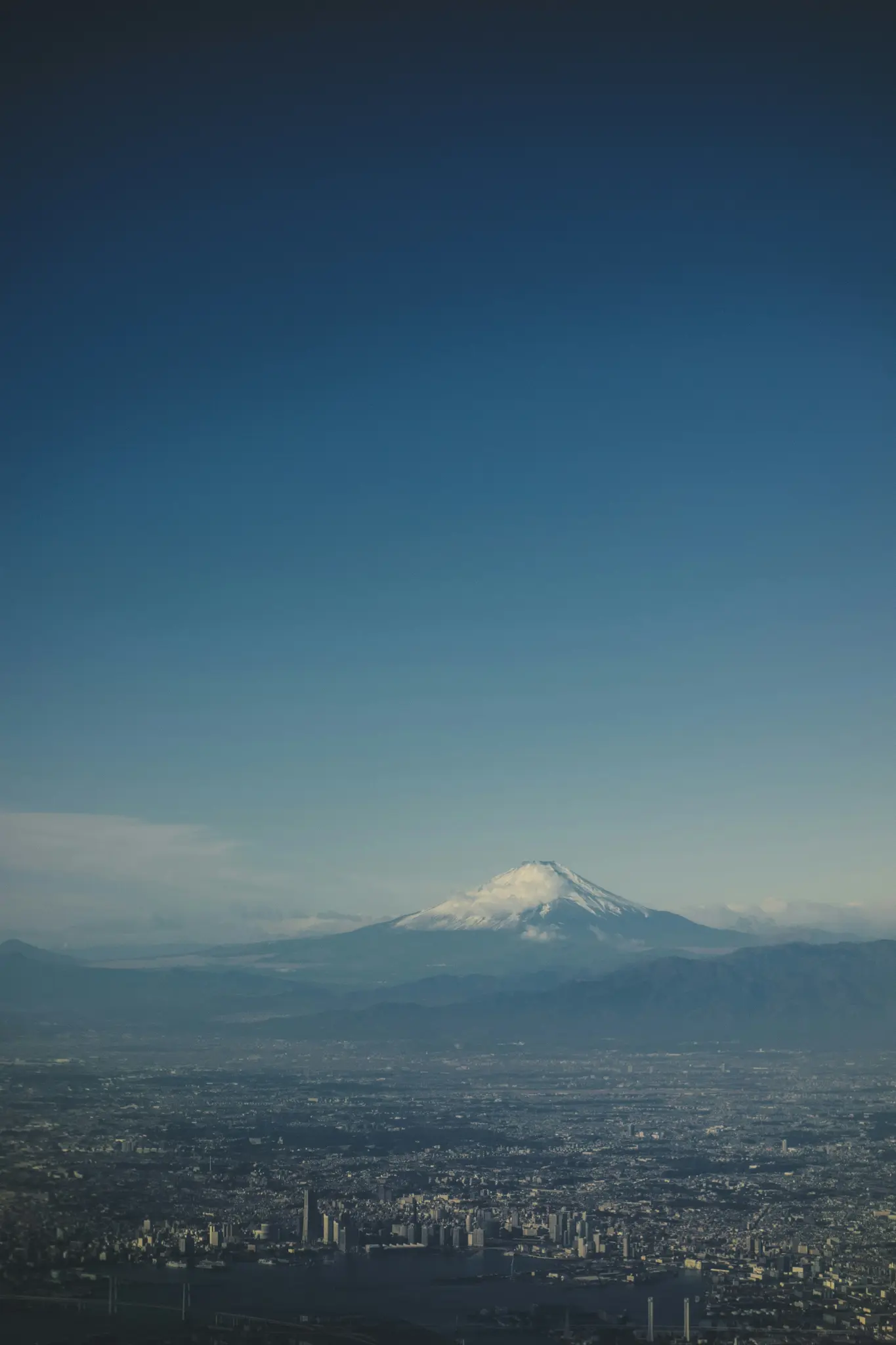 En luftudsigt over Fuji, hvis top er dækket af sne, der rejser sig over en vidstrakt by med talrige bygninger og en klar blå
