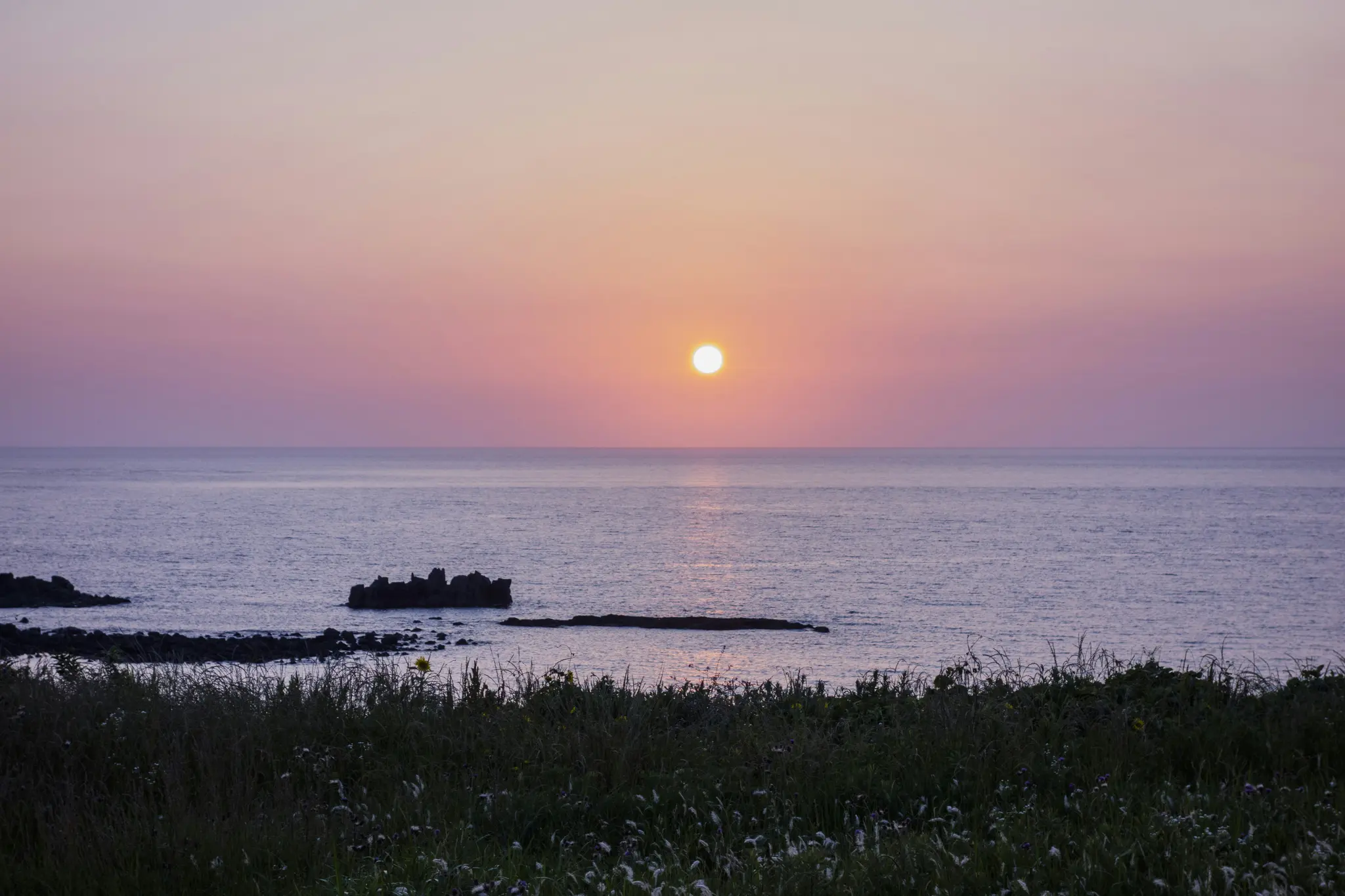 En levende solnedgang over et roligt hav, med en himmelgradient fra orange til pink til lilla, med mørke klippeformationer