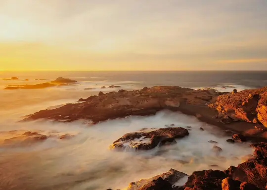 Uno scatto a lunga esposizione di una costa rocciosa al tramonto, con l'oceano che appare liscio e nebbioso mentre le onde si