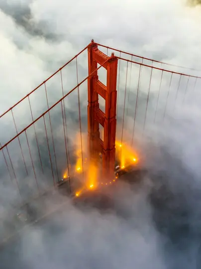 Pohled z ptačí perspektivy na horní část červeno-oranžové věže mostu Golden Gate, zahalenou hustou bílou a šedou mlhou.