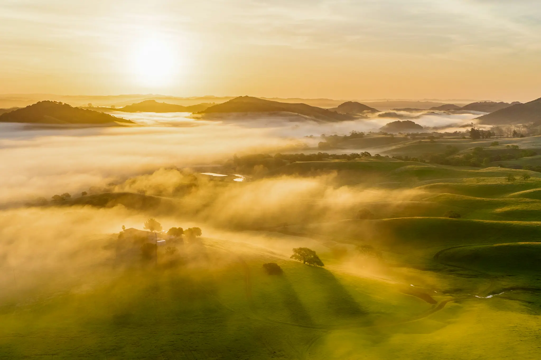 Een luchtfoto van het landschap toont een zonsopgang boven glooiende groene heuvels, gedeeltelijk bedekt door dichte, gouden