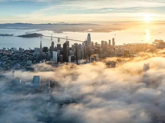 En flygvy över San Franciscos skyline vid soluppgången, med dimma som delvis skymmer byggnaderna och Bay Bridge.