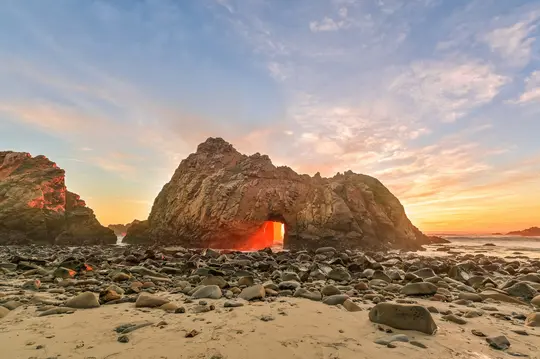 Een levendige zonsondergang verlicht een natuurlijke boog in een grote rotsformatie op een zandstrand bezaaid met gladde