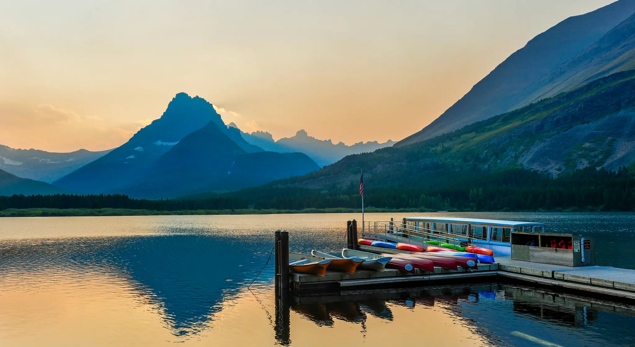 A tranquil lake reflects the warm orange and yellow hues of a golden hour sky, with dark blue mountains in the background.…