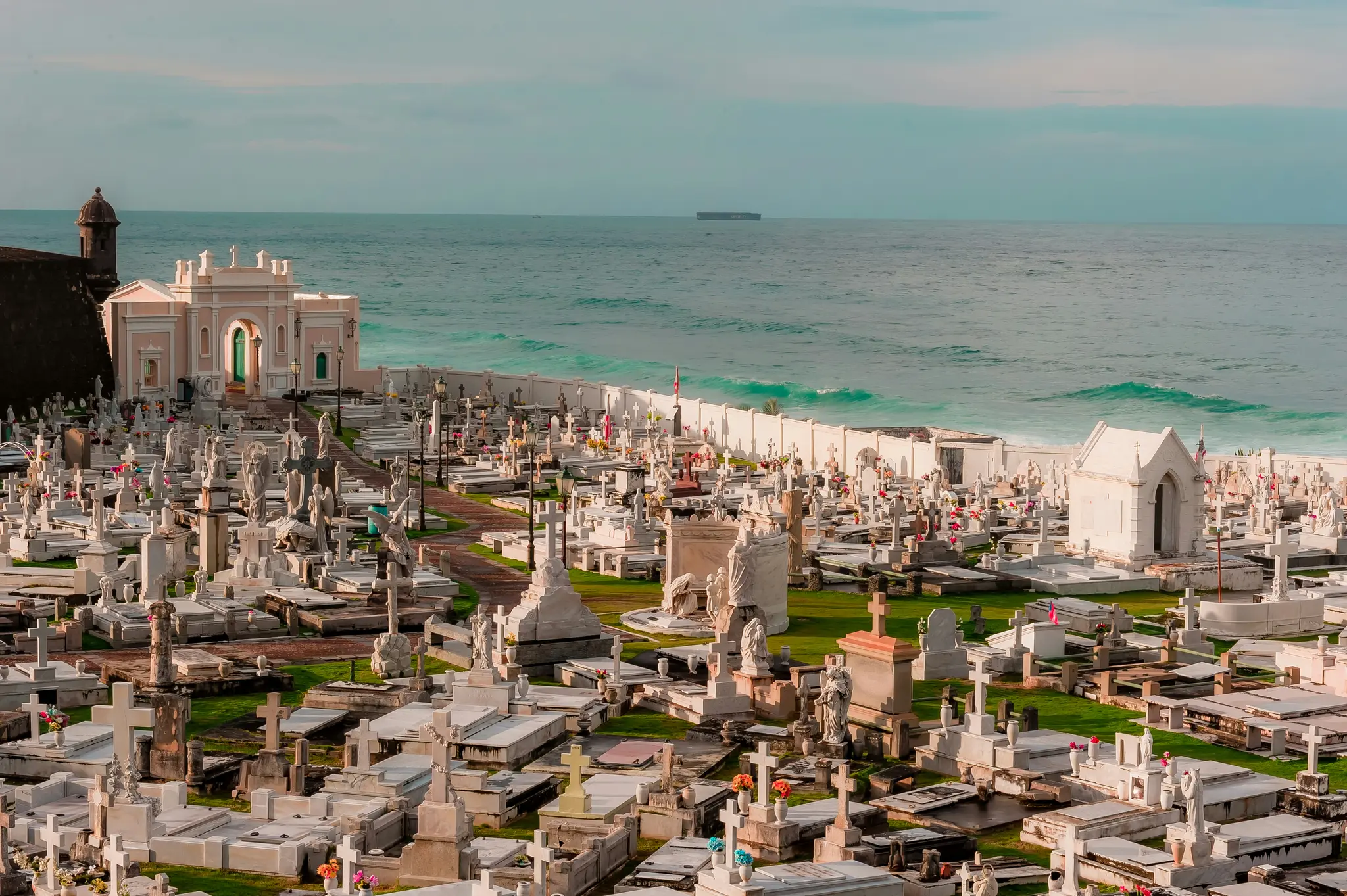 Una vista elevata di un vasto cimitero con innumerevoli tombe e monumenti bianchi, delimitato da un muro bianco e dall'oceano