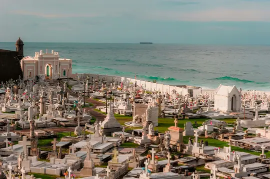 Vue aérienne d'un vaste cimetière avec d'innombrables tombes et monuments blancs, bordé par un mur blanc et l'océan bleu.