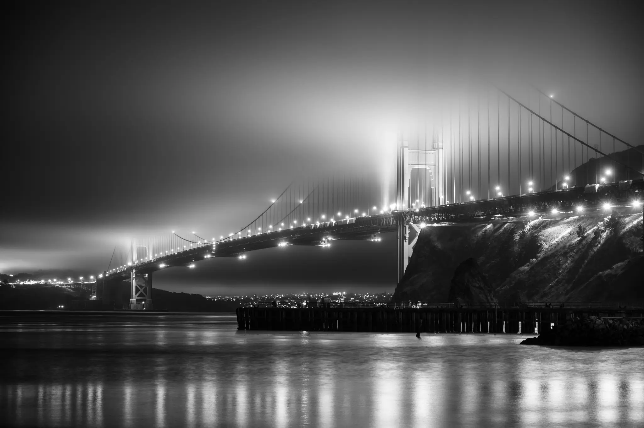 En svartvit lång exponering av Golden Gate Bridge på natten, med dess ljus som lyser genom en tjock dimma. Ljusen…