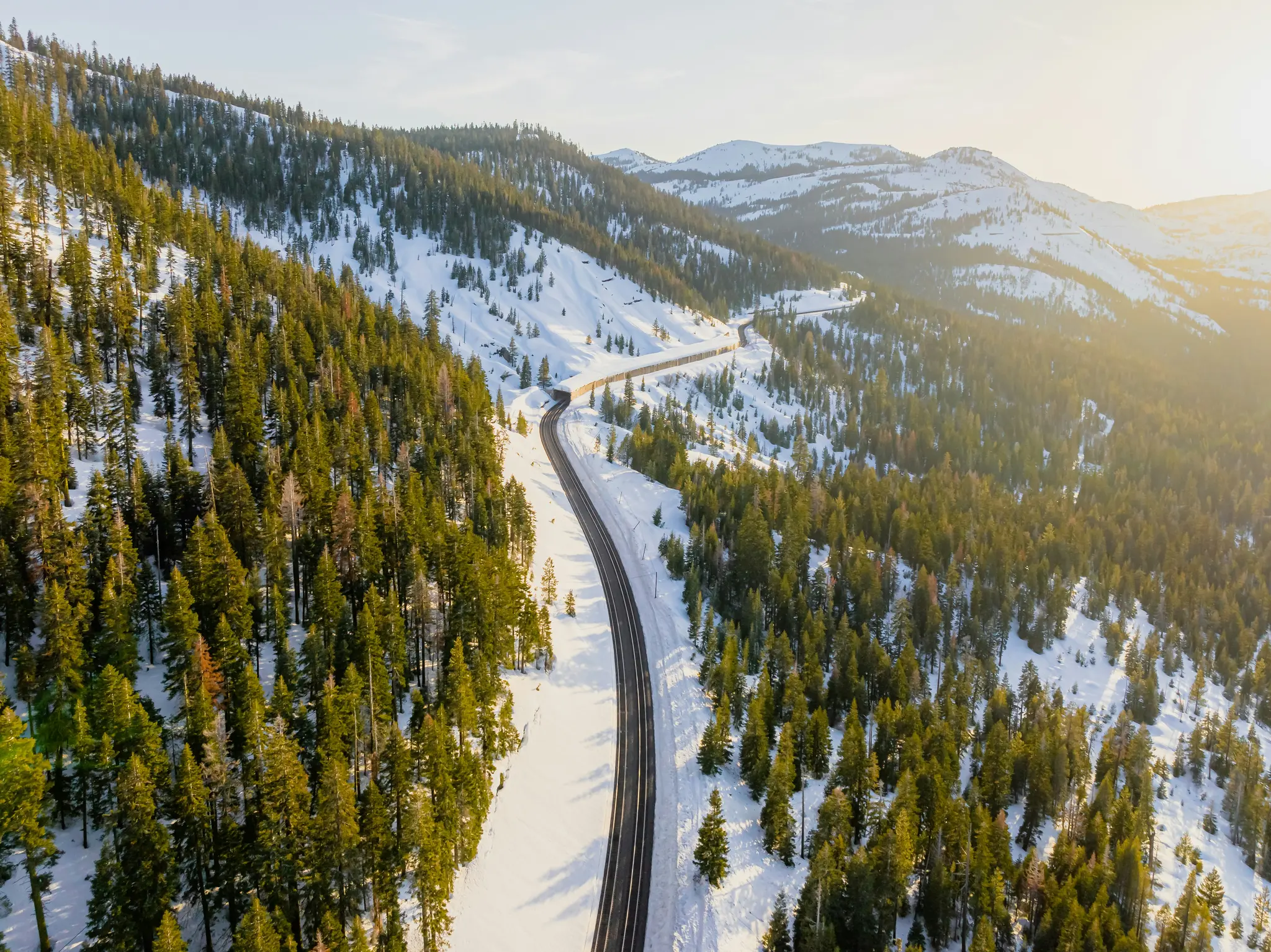 En flygvy visar en mörk, slingrande väg som skär genom ett snötäckt bergslandskap fyllt med gröna tallar.