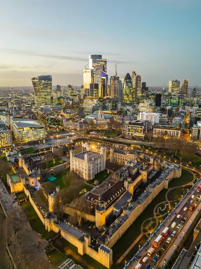 Pohled z ptačí perspektivy na Tower of London s osvětlenými hradbami a budovami na pozadí moderní londýnské panoramy.