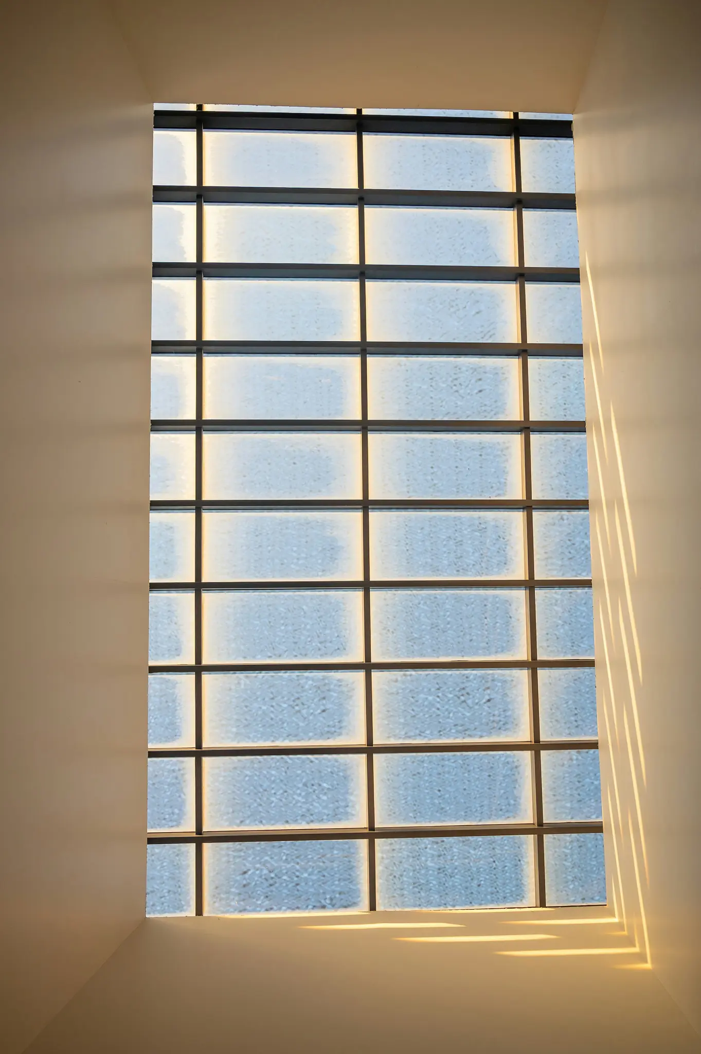Een hoog, rechthoekig raam met horizontale en verticale spijlen. Zonlicht werpt lange, parallelle schaduwen aan de