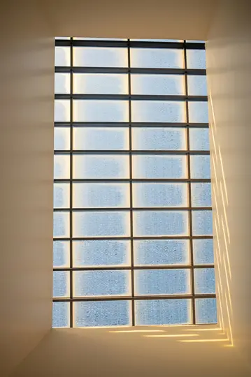 Una ventana alta y rectangular con barras horizontales y verticales. La luz del sol proyecta sombras largas y paralelas a la