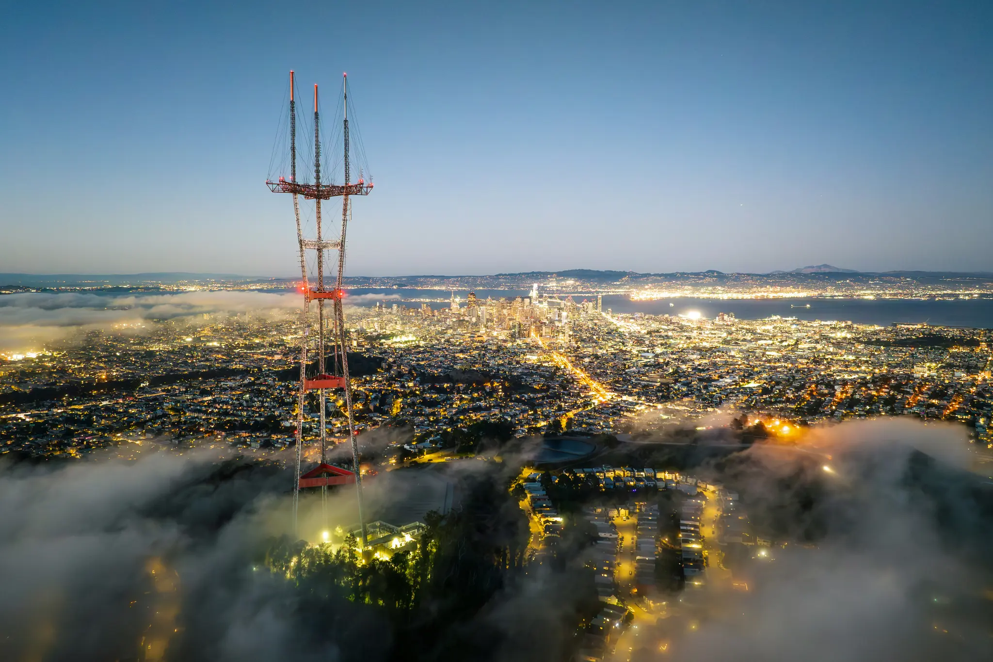 Une vue aérienne d'une ville au crépuscule, avec une haute tour de transmission au premier plan et une couche de brouillard