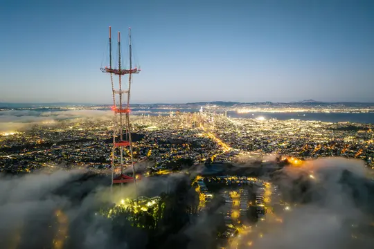 Een luchtfoto van een stad bij schemering, met een hoge zendmast op de voorgrond en een deels bedekkende mistlaag…