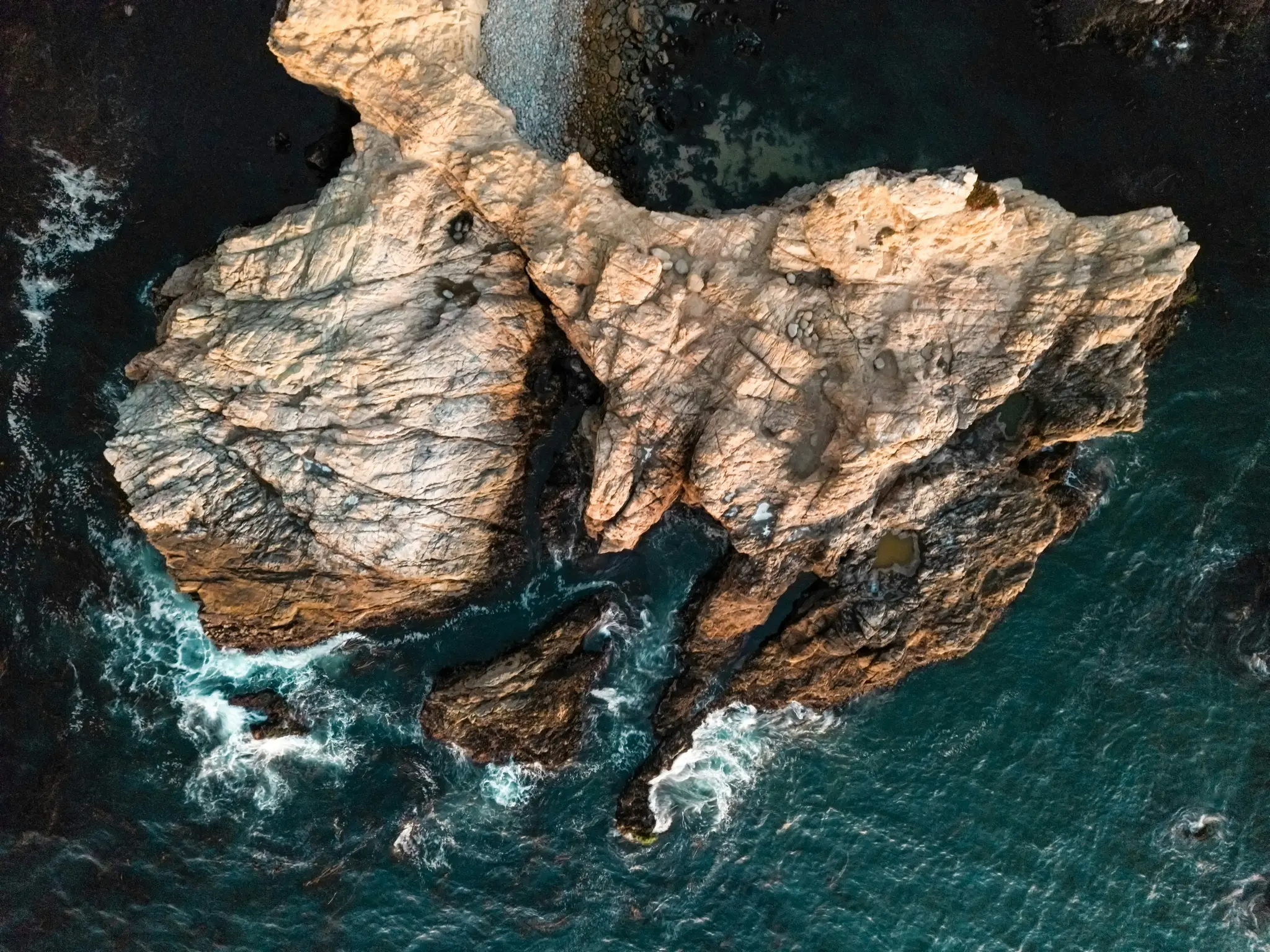Une photo aérienne montre une côte accidentée avec des rochers brun clair et brun foncé rencontrant de l'eau de mer bleu