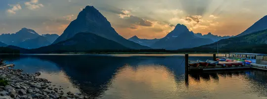 Een panoramisch landschap van een rustig meer dat een zonsonderganghemel met zonnestralen en donkere bergen weerspiegelt. Een