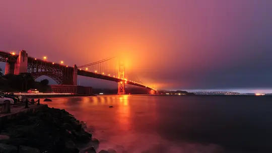 Una toma de larga exposición del puente Golden Gate por la noche, brillando de color naranja a través de la niebla, con cielo