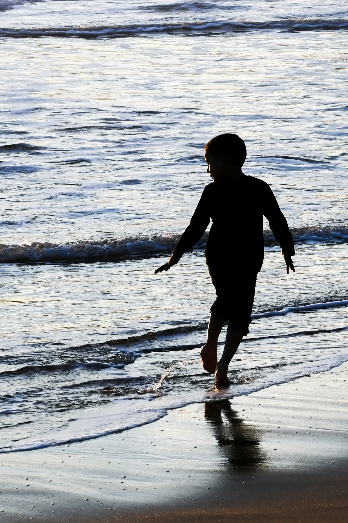 De silhouet van een kind loopt op blote voeten in het ondiepe oceaanwater tijdens het gouden uur, met zachtjes brekende