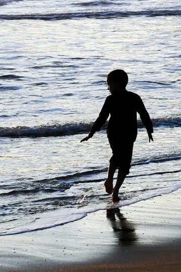 La silueta de un niño camina descalzo en el agua poco profunda del océano al atardecer, con olas rompiendo suavemente.