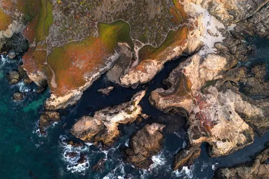 O vedere aeriană a unei coaste accidentate. Oceanul, în nuanțe de albastru profund și turcoaz, întâlnește stânci stâncoase și