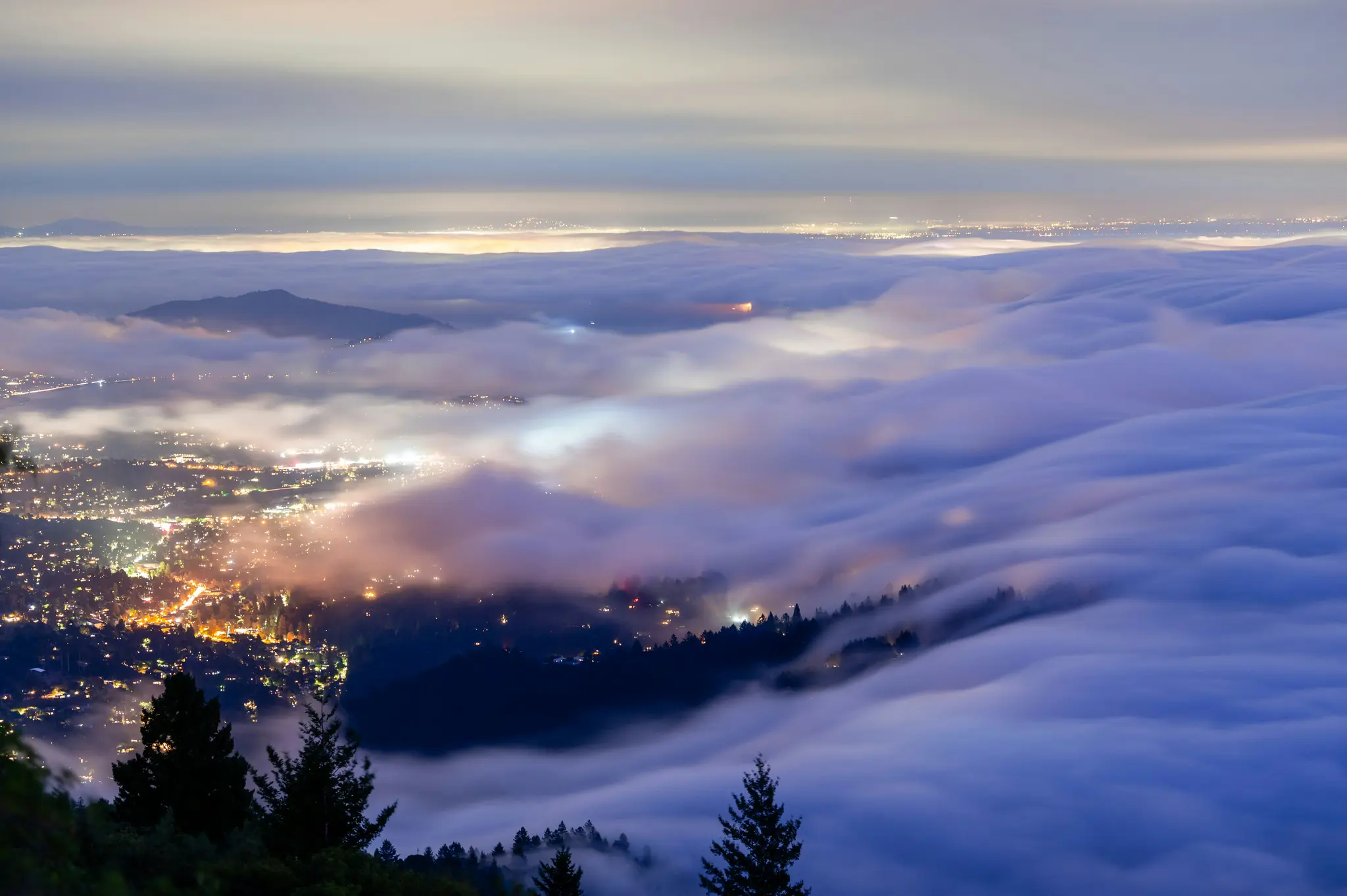 An aerial view of a city at night, with bright orange and yellow lights peeking through a thick, undulating layer of…