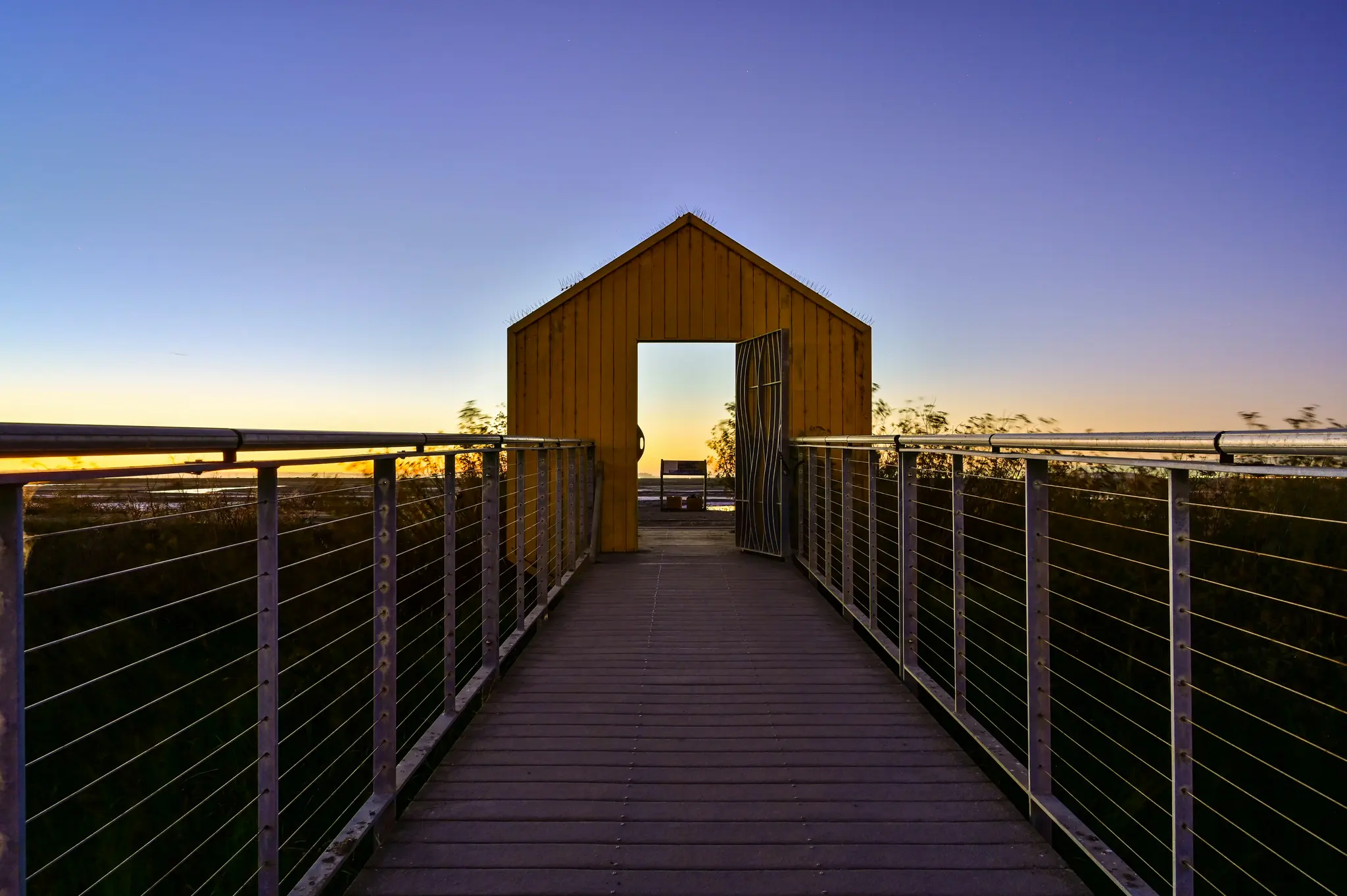 Een houten promenade met metalen leuningen leidt rechtstreeks naar een klein, geel, huisvormig gebouw met een open deur.