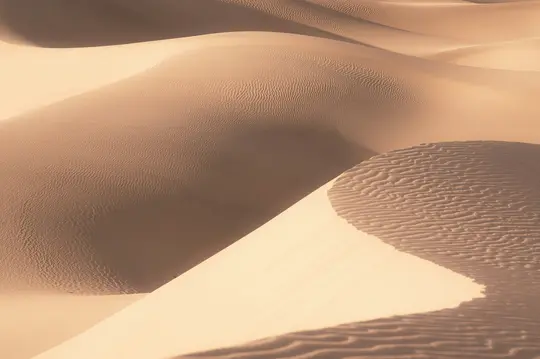Una vista ravvicinata e astratta di dune di sabbia. Le dune sono beige chiaro e marroni, con ombre morbide che ne evidenziano