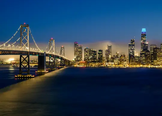 O fotografie cu expunere lungă a podului Bay Bridge din San Francisco iluminat pe timp de noapte, cu orizontul orașului