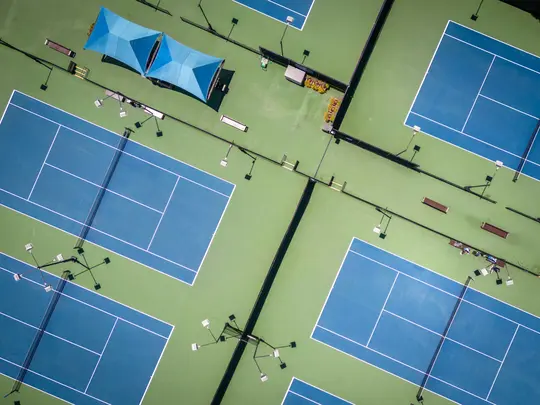 Une vue aérienne, de dessus, de plusieurs courts de tennis bleus avec des lignes blanches, entourés de verdure. Deux zones
