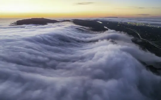 Una vista aérea muestra una vasta extensión de niebla blanca y gris que cubre un paisaje, con colinas oscuras y luces de la