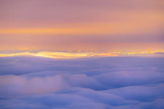Un paisaje aéreo muestra una gruesa capa de niebla de color azul púrpura que cubre la mitad inferior, con luces de la ciudad