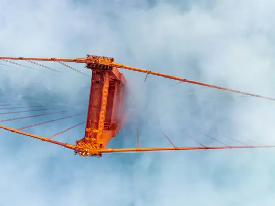 Una toma hacia arriba de una torre de puente naranja, con sus cables extendiéndose hacia afuera, parcialmente envuelta en