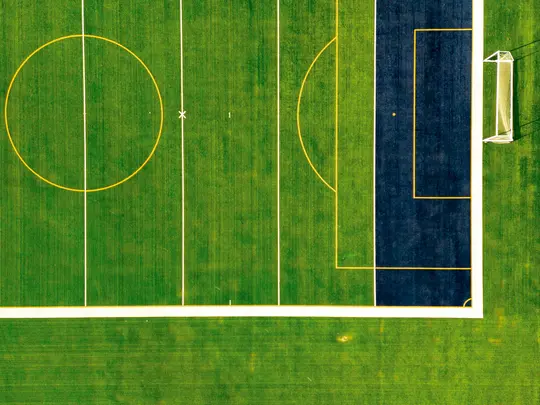 Una vista cenital de un campo deportivo de color verde vibrante con líneas blancas y amarillas, y un área marcada de color