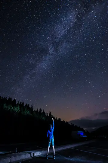 Une personne en veste bleue et short se tient sur un parking sombre, pointant vers un ciel rempli d'innombrables étoiles et