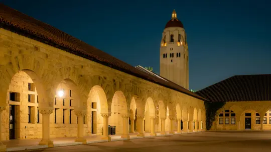 Une longue arcade en pierre avec de nombreuses arches, illuminée par des lumières chaudes la nuit, menant vers une haute tour