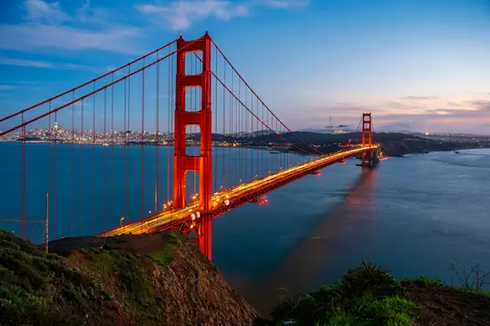Fotografie cu expunere lungă a podului Golden Gate la apus, cu turnurile și cablurile sale roșii iluminate pe un fundal
