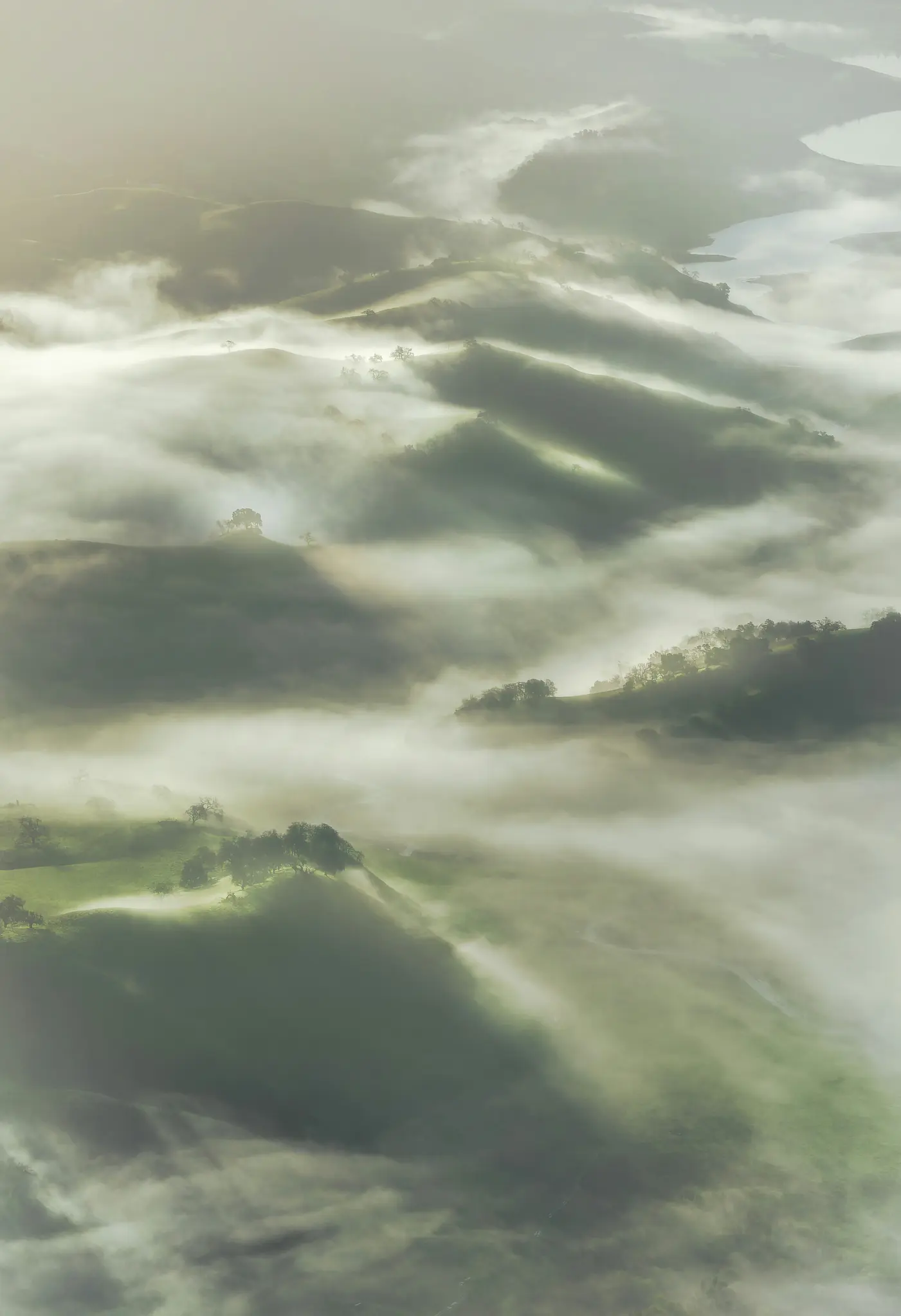 An aerial photograph shows a landscape of rolling green hills partially covered by a thick, white fog. A body of water is…