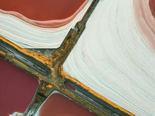 O vedere aeriană abstractă a iazurilor de evaporare a sării, arătând zone de roșu intens, roz pal și alb cu linii texturate.